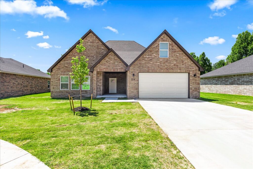 A brick home with a white two-car garage sits on a lot with a long driveway. A blue sky with clouds and a green lawn frame the photo.