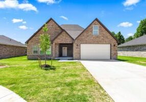 A brick home with a white two-car garage sits on a lot with a long driveway. A blue sky with clouds and a green lawn frame the photo.