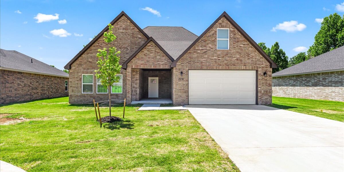 A brick home with a white two-car garage sits on a lot with a long driveway. A blue sky with clouds and a green lawn frame the photo.